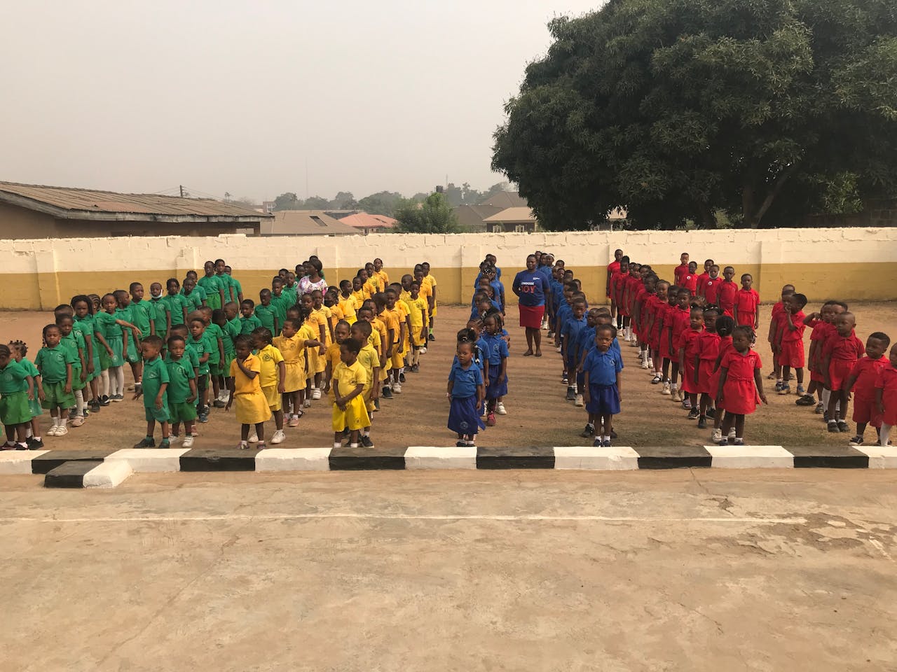 Children in colorful uniforms stand in formation on a school ground for assembly.